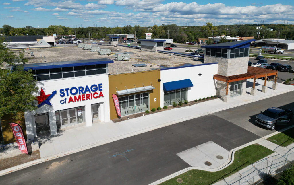 Aerial view of a single‑story commercial building with a “Storage of America” sign on the left storefront, blue awnings, landscaped planters, and a paved road in front.